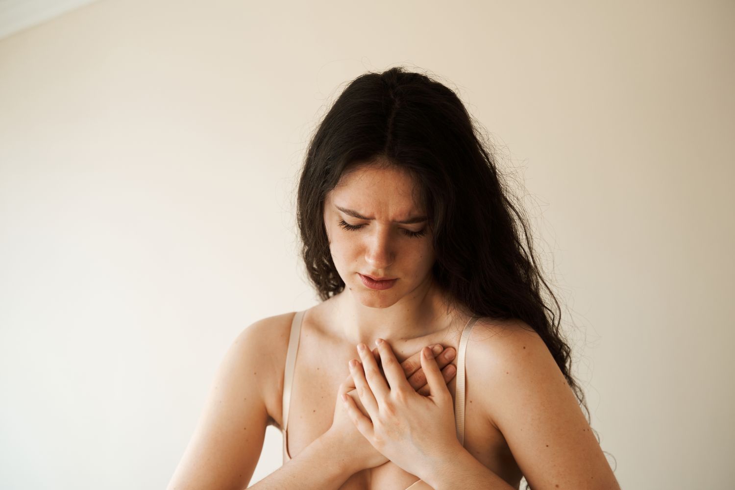 A imagem mostra uma mulher com expressão de dor, pressionando o peito com as mãos. O cenário neutro destaca o desconforto, possivelmente associado a ansiedade causa dor na mama.