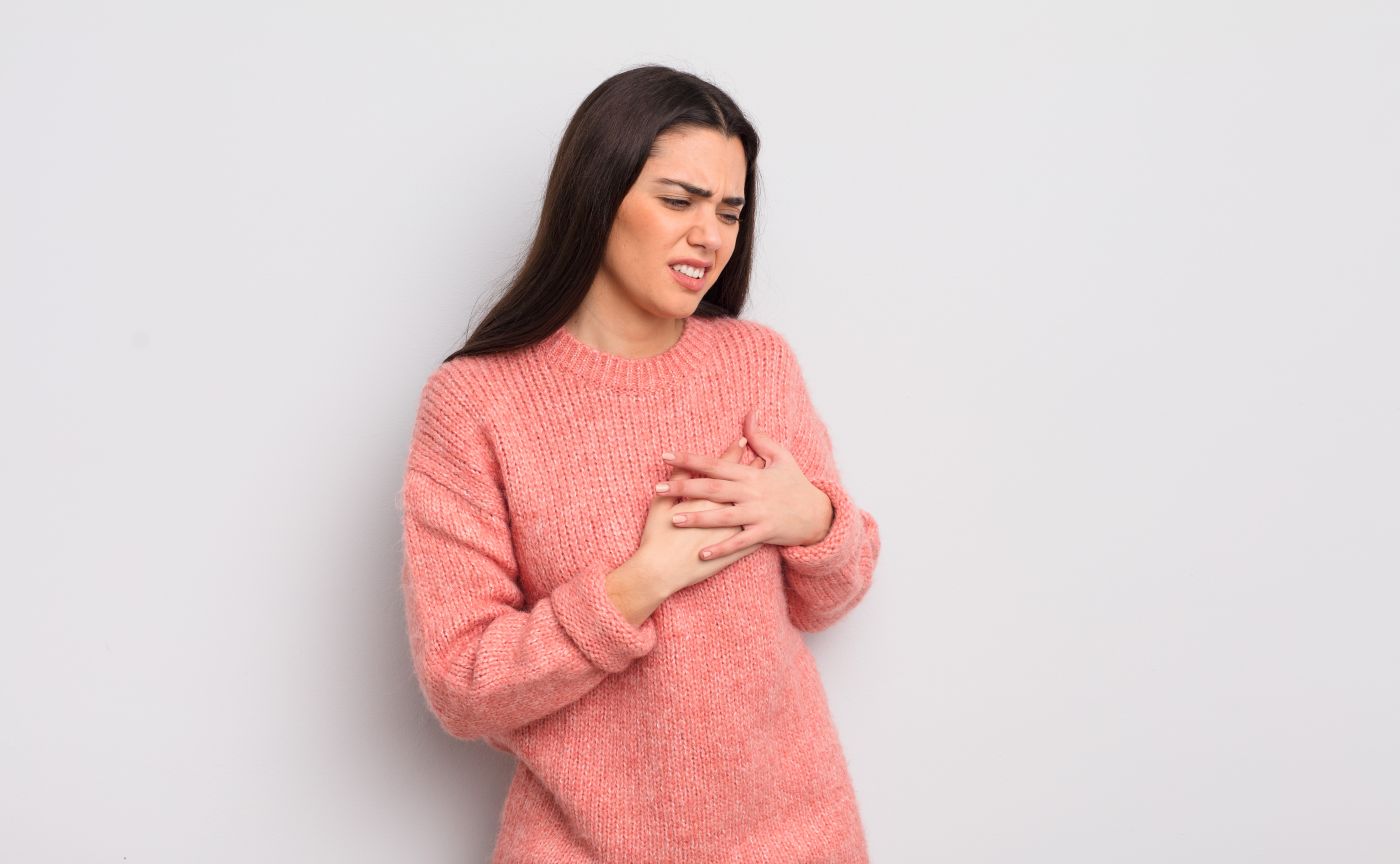 A fotografia de estúdio em fundo cinza claro mostra uma jovem com longos cabelos castanhos expressando dor ou desconforto emocional e físico, como algo provocado por nódulo na mama que dói ao apertar.