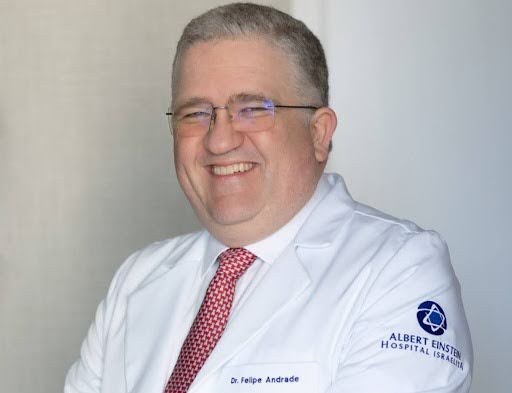 O retrato destaca o Dr. Felipe Andrade, médico mastologista, que sorri e veste jaleco branco com gravata vermelha, transmitindo uma imagem profissional