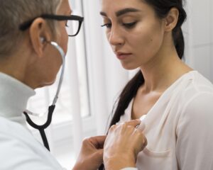 Mulher sendo examinada por médica com estetoscópio na região do peito durante avaliação clínica relacionada aos cuidados após biópsia da mama.