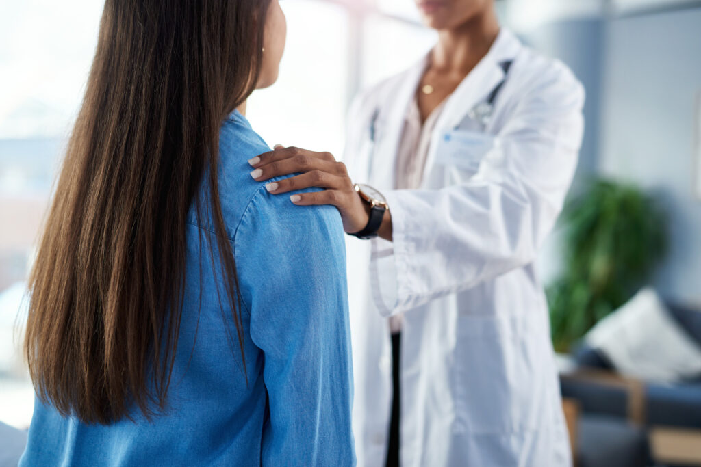 Médica de jaleco branco apoiando a mão no ombro de paciente durante consulta médica, transmitindo acolhimento e confiança em ambiente clínico especializado em saúde da mulher.