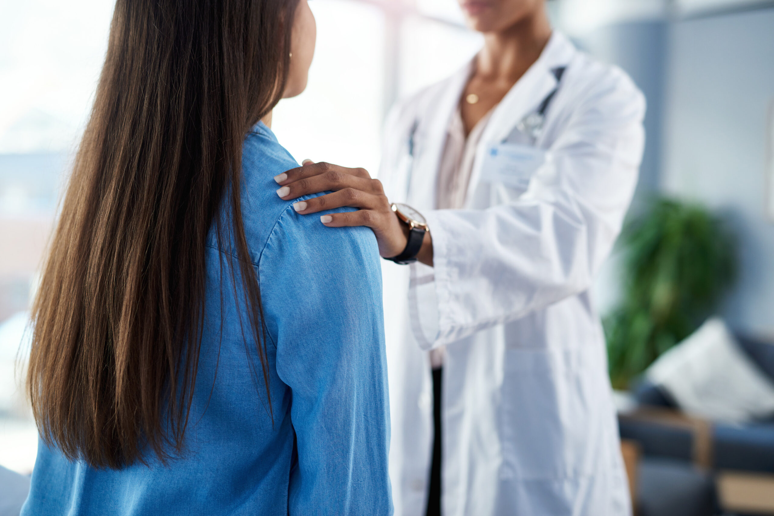 Médica de jaleco branco apoiando a mão no ombro de paciente durante consulta médica, transmitindo acolhimento e confiança em ambiente clínico especializado em saúde da mulher.