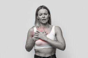 Mulher com expressão de dor segura o peito com as duas mãos, com área destacada em vermelho indicando sensibilidade ou desconforto na mama.
