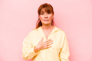Mulher com a mão sobre o peito em fundo rosa, representando atenção à saúde da mama.