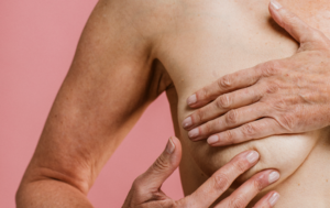 Mulher realiza autoexame das mamas, apalpando uma das mamas com as mãos contra um fundo rosa, em imagem relacionada à avaliação da saúde mamária.