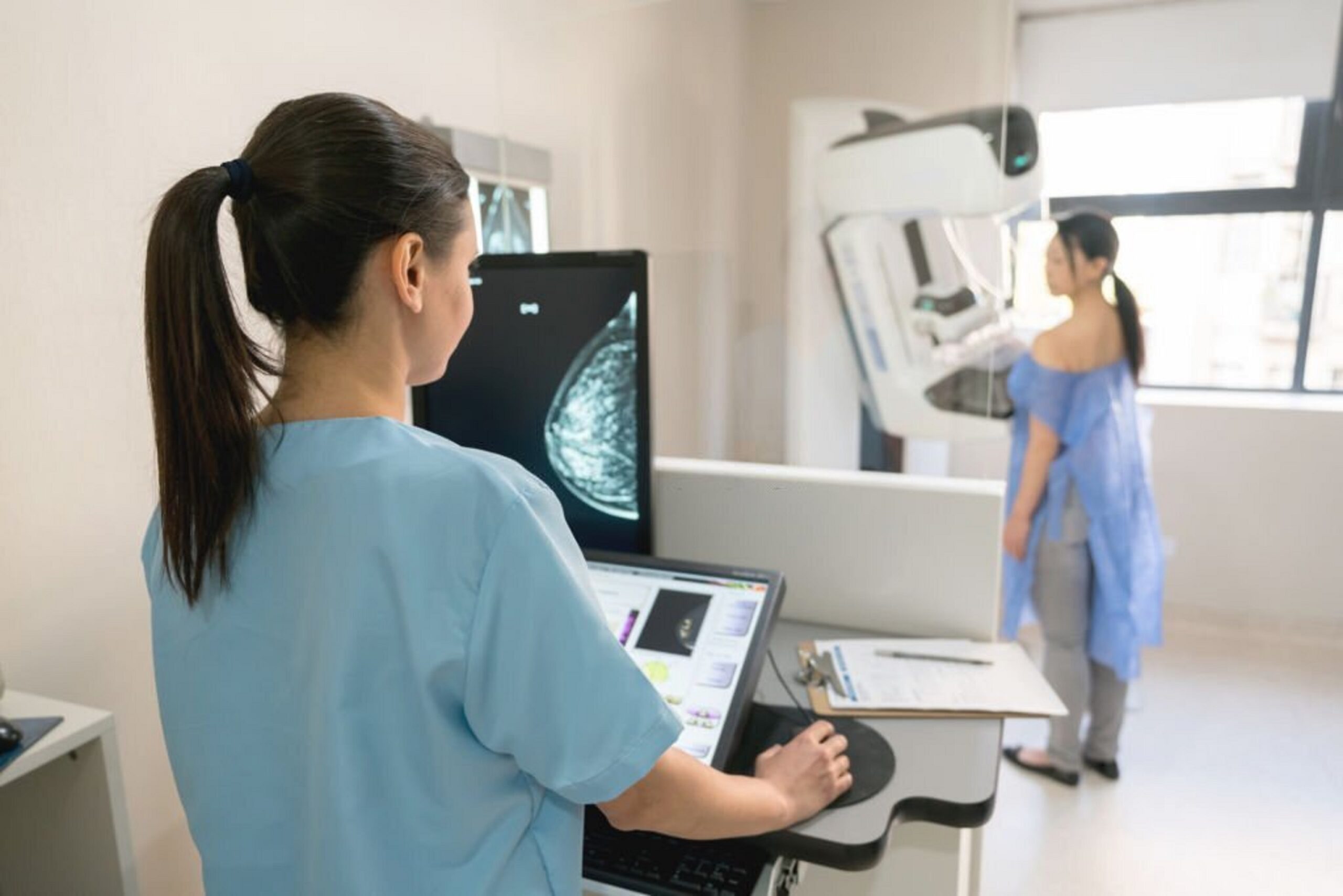 Profissional de saúde opera equipamento de mamografia enquanto paciente se posiciona para a realização do exame em uma clínica de diagnóstico por imagem.