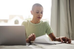 Mulher com cabeça raspada lendo um documento em casa, ao lado de um notebook, representando o momento de análise de resultados médicos como BI-RADS 3.