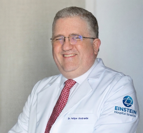 Retrato profissional de um homem sorrindo, usando óculos, jaleco branco, camisa clara e gravata vermelha. Ele está sentado diante de um fundo cinza-claro, com o logotipo do Einstein Hospital Israelita visível na manga do jaleco.