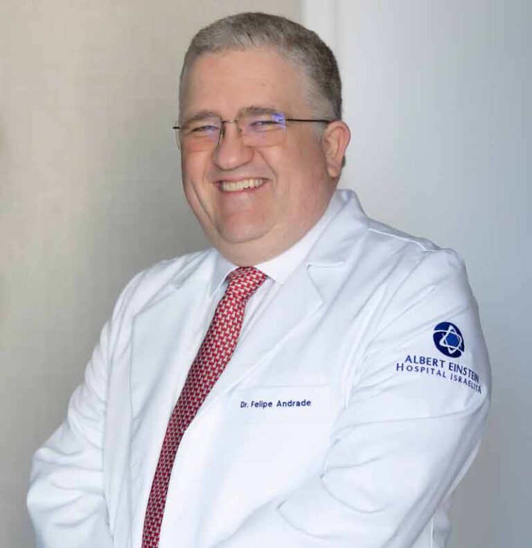 Retrato institucional de médico sorrindo, com cabelos grisalhos, óculos, jaleco branco e gravata vermelha. No jaleco aparece o nome “Dr. Felipe Andrade”.
