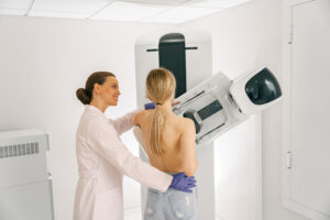 Profissional de saúde acompanha uma mulher durante exame de mamografia, posicionando-a diante do equipamento em uma sala de diagnóstico por imagem.